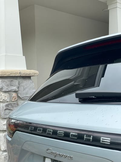 The image shows the rear of a light gray Porsche Cayenne parked near a stone and white-painted brick wall. The tailgate is open, reflecting the exterior of a house. Water droplets are visible on the car's surface, suggesting a recent rainfall. "PORSCHE" is spelled out vertically on the black trim surrounding the taillight, with "Cayenne" written in cursive on the right. The photograph appears to be taken outdoors in Windermere, United States.