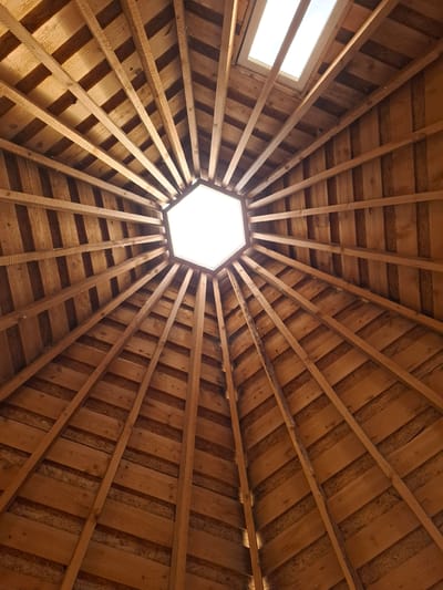 The image captures an upward view of an interior wooden ceiling or roof structure, indicative of a circular or polygonal building, set in Ludorvai, Russia. The central feature is a bright, hexagonal skylight, acting as a primary light source. Numerous wooden beams or rafters extend radially outwards from this central opening, forming a spoke-like pattern across the ceiling.
These beams are made of natural, unpainted wood, revealing visible grain and knots, suggesting a rustic yet robust construction. Between the radial beams, the ceiling surface is covered with horizontal wooden planks, adding texture and depth to the overhead view. Towards the upper left periphery of the frame, a secondary, rectangular skylight or window is visible, also admitting a significant amount of bright, natural light.
The strong illumination from both skylights indicates it is daytime, likely under clear weather conditions, as sunlight appears unobstructed. The light highlights the warm, earthy tones of the wood and creates subtle shadows that emphasize the architectural details. There are no people, activities, or visible text in the image. The scene highlights the intricate wooden craftsmanship and the natural light flowing into the space.
