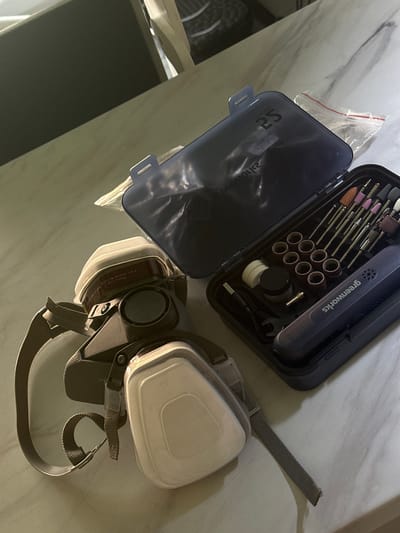 An overhead, tilted shot displays a rotary tool kit and a respirator mask laid out on a light-colored, possibly marbled, surface in an indoor setting in Kuda, Russia. The lighting appears to be artificial.
The primary objects are an open, dark blue or black case containing a rotary tool and its accessories, and a multi-part respirator mask. The rotary tool, branded "greenworks," is visible inside its organized case, surrounded by various attachments including grinding stones, sanding drums, and other bits. A small wrench, a charging cable, and what appears to be a small jar of polishing compound or similar accessory are also neatly arranged within the case. Reflective text inside the case lid shows "2S" and what appears to be a reversed "greenworks" logo.
Adjacent to the tool kit is a respirator mask, positioned with its filter cartridges facing upwards. The mask is dark grey and light beige, featuring two large, rectangular filter cartridges attached to the main body. Visible text on one of the cartridges includes "Plastic filter," "P-1," "for industrial air supplies only," and instructions like "original No, replace," and "Replace filters regularly." The mask has several adjustable straps, one of which extends across the frame. A small, clear plastic bag with a red zip-lock is situated between the tool case and the respirator.
The arrangement suggests the items are either recently used, being prepared for a task involving fine detail work, or stored. No people are visible, and no specific activity is taking place.