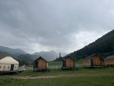 This is a landscape photograph taken in Chelpek, Kyrgyzstan, likely in the late afternoon given the overcast sky. The scene depicts a rural glamping or eco-resort with several small wooden cabins and a traditional yurt. The cabins are uniformly designed, elevated on stilts, and feature dark metal roofs and doorways. A white picket fence runs horizontally across the middle ground, separating the cabins and yurt from the natural landscape beyond.

In the background, rolling green hills transition into rugged, mist-shrouded mountains, suggesting a mountainous or high-altitude region. The sky is heavily clouded, with a uniform grey tone, indicating a possibility of rain or generally gloomy weather. The ground in the foreground is covered with green grass, with a dirt path winding through it. A few cars are visible parked in the distance behind the yurt, near some trees and utility poles.

The overall mood of the image is serene and slightly melancholic, due to the weather and the isolated setting. There are no people visible in the frame, giving the impression of a quiet and peaceful retreat. The architecture of the yurt, with its distinctive patterned fabric and dark accents, hints at the cultural context of the location. The image captures a harmonious blend of traditional and modern accommodation within a natural, mountainous environment.
