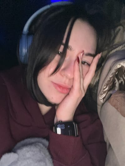 A young woman poses indoors wearing a maroon hoodie. Part of her face is obscured by her hand. She is also wearing an Apple Watch with a leather band. The timestamp visible on the watch display reads 07:03. A headphone is visible on the left side of the image. She has dark hair. The background includes a textured blanket.