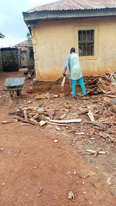 A person is seen from behind, wearing a light blue overshirt and bright teal pants, working with a pile of cut wood. They are holding a piece of wood or similar material in their hands, appearing to be moving or sorting it. The ground is a reddish-brown dirt, littered with many pieces of wood of various sizes and shapes, some with bark still attached. To the left of the person, there is a blue wheelbarrow. Behind the person, a light-colored building with a corrugated metal roof is visible. The building has a small window with bars and appears to have some writing or graffiti on its exterior wall. In the background, other structures, including a brick wall and another building with a similar corrugated roof, can be seen. There are also indications of vegetation in the distance and a couple of chickens are visible near the brick wall. The sky is overcast, suggesting a cloudy day. The scene appears to be an outdoor, possibly rural, setting in Kagarko South, Nigeria, where wood is being processed or collected. The overall mood seems practical and industrious, focused on the task at hand. The writing on the wall of the building is not clearly legible.