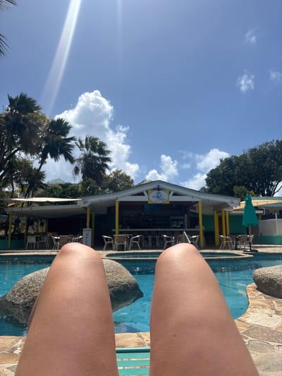 The image depicts a sunlit tropical scene in Olde Yard Village, British Virgin Islands, captured from a first-person, low-angle perspective. The foreground is dominated by the tanned thighs and knees of an individual, presumably lying on a lounge chair, a portion of which is visible as a teal surface at the bottom edge.
Immediately beyond the foreground, a swimming pool with clear blue water and visible light blue tiling occupies the midground. Two large, smooth, grey boulders are partially submerged within the pool, positioned on either side of the viewer's legs.
In the background, a brightly colored, open-air building, likely a bar or restaurant, serves as the central focal point. The structure features a red main body with prominent yellow support columns and trim, topped by a white gabled roof section. A yellow sign bearing the "The Village" logo is affixed to the gable. Beneath this section, a bar counter with multiple stools is visible. To the left of the bar, a covered patio area with several light-colored tables and chairs is present; an "OPEN" sign is partially visible on a nearby post. To the right, another small seating area with similar furniture is shaded by a large, open teal umbrella.
The surrounding environment is characterized by lush tropical vegetation, including numerous palm trees with both fan-shaped and feathered fronds, and dense green trees, framing the building and extending upwards into the sky. The sky is a clear, bright blue with scattered white cumulus clouds. A prominent sun flare or lens flare originates from the upper left portion of the sky, indicating direct sunlight. No other individuals are distinctly observed.