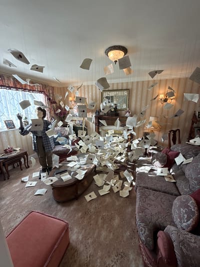 The image depicts a whimsical scene filled with countless pieces of paper, resembling letters or envelopes, suspended in the air by thin threads. The setting appears to be an indoor room, possibly a living room or a themed exhibit. A mannequin dressed in a plaid shirt and brown pants is positioned in the center, seemingly interacting with the floating papers, with one hand reaching out. The room has ornate furniture, including a large patterned sofa and a wooden table. The walls are decorated with striped wallpaper and a framed picture. A lamp with a decorative shade hangs from the ceiling, and a chandelier is visible in the background. The overall impression is one of a magical and chaotic event, reminiscent of the Harry Potter series with its iconic flying letters. The lighting suggests daytime, and there are no clear indicators of weather conditions. The scene evokes a sense of wonder and enchantment.