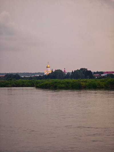 The image shows a serene, overcast scene in Kazachinskoe, Russia, likely during the evening or early morning. A calm river occupies the foreground, reflecting the muted light of the sky. The middle ground features a lush, green treeline that separates the water from a village. Prominently visible within the village is a white church with a golden dome, characteristic of Russian Orthodox architecture. To the left of the church, a smaller structure with a pink, conical roof is also present. Several residential buildings with red roofs are visible further to the right, indicating a populated area. The sky is a soft gray, with subtle variations in tone suggesting clouds. No people or immediate actions are depicted; the image captures a tranquil, static view of the landscape and settlement.