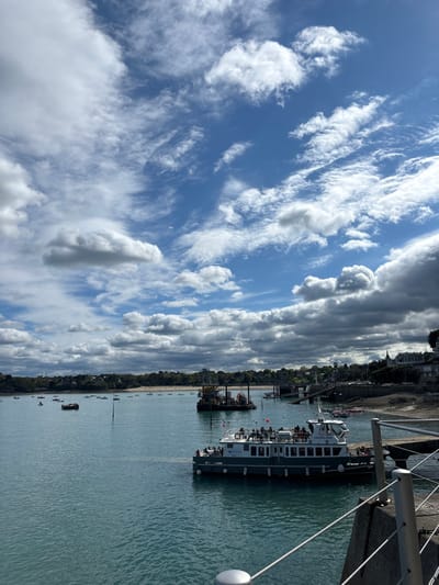 A large ferry boat is docked at the harbor in Dinard, France (latitude: 48.634, longitude: -2.049). The ferry boat is grey and white with a French flag on the stern. There are people on board the boat, and several people are standing on a dock in the background. The water is calm and blue, and the sky is a bright blue with white clouds. The view is from the edge of the harbor, looking out toward the sea. There is a small rocky beach on the right-hand side of the image and a small pier. The background shows trees and buildings, with more buildings visible in the distance. The scene appears to be on a sunny day. There is a metal cable railing in the foreground.