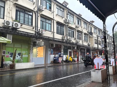 In the Xuhui District of China, on a rainy day, a street scene unfolds with buildings lining the wet pavement. The buildings are multi-story with many windows and air conditioning units visible. A row of shops runs along the ground floor, one featuring a bright green facade with a cartoon cow advertisement and the word "YOGURT". A person sits under a clear umbrella next to a potted plant outside this shop.
Further down the street, a small blue motorized tricycle is parked, partially covered with a colorful cloth. Next to it, a yellow scooter is parked, covered with a yellow tarp. A person wearing a helmet and a backpack rides a dark motorcycle in the rain, heading away from the camera. In the background, other vehicles and pedestrians with umbrellas are visible. The street is wet and reflects the lights from the surrounding environment. Planters with white textured sides are placed along the sidewalk, some with white decorative elements. The sky is overcast, indicating continued rain.