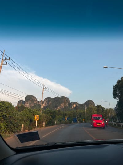 The image depicts a roadside scene in Ban Nai Sa, Thailand. A red vehicle is traveling on a road with mountainous terrain and vegetation in the background. Overhead power lines and utility poles are visible. Roadside signage is also present. The view is captured from inside a vehicle.