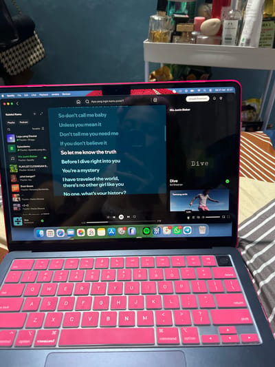 The image displays a close-up of a personal computing setup, likely in a bedroom in Magelang, Indonesia. A tablet or iPad, encased in a pink cover, rests on a patterned bedsheet, positioned vertically. It is displaying the Spotify music streaming application, showing the lyrics for the song "Dive" by Ed Sheeran. The visible lyrics include phrases like "So don't call me baby Unless you mean it Don't tell me you need me If you don't believe it So let me know the truth Before I dive right into you You're a mystery I have traveled the world, there's no other girl like you No one. what's your history?". The song is at the 1:12 mark out of 3:58.
Next to the tablet, a laptop with a prominent pink keyboard cover is also visible, suggesting a cohesive aesthetic choice. The top bar of the tablet indicates the current time as "Sel 21 Apr 22.31" (Tuesday, April 21st, 10:31 PM). The Spotify interface is largely in Indonesian, with text like "Apa yang ingin kamu putar?", "Jelajahi Premium", "Mix Justin Bieber", and various playlist titles including "PLAYLIST CLOSINGAN B...", "jahat banget?", "Brain Boost", and "<3<3". The tablet's dock shows app icons for Settings, App Store, Photos, Messages, Safari, Mail, Calendar, Reminders, Files, WhatsApp (with 127 unread messages), "Tut on", and Spotify.
In the background, slightly out of focus, there is a shelf holding various bottles, some labeled "THE BODY SHOP," "KALIGRAFI," and "RAI," suggesting personal care products or perfumes. The overall scene depicts a relaxed, private setting where someone is enjoying music.