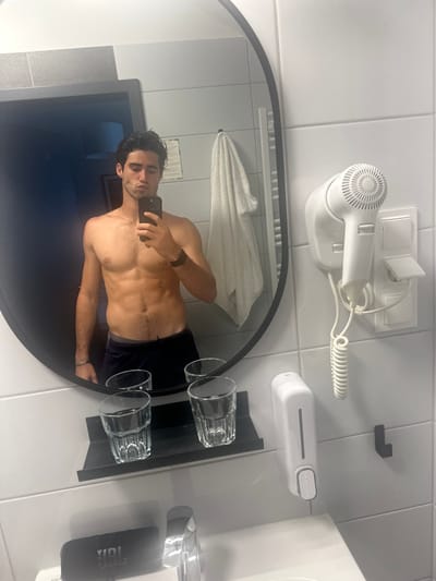 The image displays a male individual taking a mirror selfie within a modern bathroom, likely situated in a hotel or guesthouse in Giebułtów, Poland. The subject, a male with dark, damp hair and an athletic build showcasing defined abdominal muscles, is shirtless and wearing dark shorts or swim trunks. He holds a black smartphone in his right hand, positioned at chest level, capturing his reflection in a large, oval mirror with a black frame. His expression features slightly pursed lips.
The bathroom environment consists of white tiled walls. A white porcelain sink with a chrome faucet is visible at the bottom of the frame, beneath the mirror. Directly below the mirror, a black wall-mounted shelf holds two clear glass tumblers with patterned textures. A black JBL portable speaker is placed on the sink counter to the left of the faucet. To the right of the mirror, a white wall-mounted hairdryer with a coiled cord is affixed to the wall, alongside a white wall-mounted soap dispenser and multiple white electrical switches or outlets. A small black hook is also present. The mirror's reflection to the subject's left reveals a white towel hanging on a wall-mounted rail and a piece of paper with text affixed to the wall. Further in the reflection, a dark room or hallway is visible through an open doorway, with a light fixture faintly discernible. The scene is illuminated by bright, even lighting.