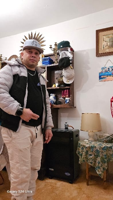 The image shows a male individual standing indoors, within what appears to be a private residence in the city of Real Sitio de San Ildefonso, Spain. The man, of apparent adult age, is positioned centrally, facing the camera with a neutral expression. He is wearing a light grey baseball cap with a white "G" emblem, an unzipped light grey and black puffer jacket over a black t-shirt, and light-colored sweatpants. A silver-colored chain necklace is visible, as is a dark smartwatch on his left wrist. His left hand is partially curled and positioned near his waist.
The background consists of a white wall adorned with various decorative and functional items. To the man's left, a golden sunburst-shaped wall ornament is partially visible. To his right, a wooden wall shelf unit holds multiple objects, including a brass candelabra, blue items, several baseball caps, a decorative teapot, bottles, and a blue and white patterned vase. Further to the right, a framed picture depicting a building scene hangs on the wall above a decorative plaque with a beach motif. A small wooden stool, draped with a blue and white floral tablecloth, supports a table lamp with a beige shade and a white base, alongside a clear glass bowl containing miscellaneous items. A black, rectangular portable gas heater on wheels, bearing the brand "ORBIEA," is situated on the tiled light brown floor to the man's right. The lower-left corner of the image contains a "Galaxy S24 Ultra" watermark.