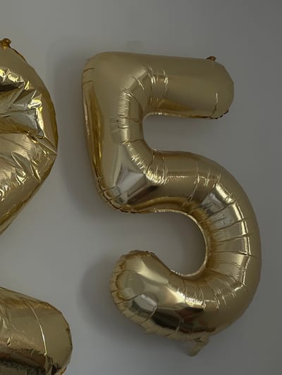 The image shows two large, gold Mylar balloons that form the numbers "2" and "5". The balloons are inflated and appear to be attached to a white wall. The lighting suggests an indoor environment. The balloons are positioned to the left and center of the frame, with the number "2" partially visible on the left and the number "5" prominently displayed in the center. There are no people or other objects visible in the image. The location is not specified in the image.