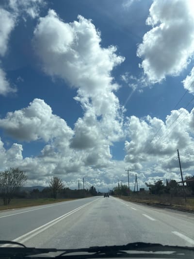 The image presents a forward-facing view from inside a vehicle on a paved two-lane road within the Prosotsani Municipal Unit, Greece. The road features a double solid white line separating the lanes and solid white lines marking the shoulders, extending towards the horizon. A dark-colored passenger car is visible in the distance, traveling in the same direction in the right lane. The foreground includes the upper portion of a vehicle's dashboard and windshield wipers. The sky above is bright blue, partially covered by numerous large, white cumulus clouds, with visible sun glare in the upper right. To the left of the road, sparse trees and dry vegetation are present, with distant hills visible on the horizon. The right side of the road features multiple wooden utility poles with overhead lines, dry ground cover, and a partially obscured red circular sign. A building with a reddish roof structure is partially visible further along the right side of the road. No individuals are depicted.