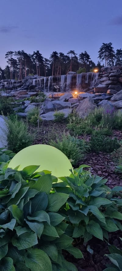 The image depicts a man-made waterfall and rock garden landscape at dusk in Shulgino, Russia. A large, glowing, spherical light fixture, casting a lime-green illumination, is situated in the foreground amidst lush green hosta plants. Behind the foliage, a multi-tiered rock formation forms the backdrop for a waterfall cascading down. Several lights are strategically placed among the rocks, illuminating the falling water and the surrounding stone structures. Pine trees line the horizon above the waterfall. The sky is a deep blue, indicating twilight. There are no people or animals visible in the scene.
