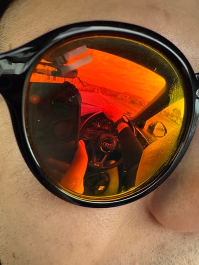 The image displays a close-up view of a person wearing black-framed sunglasses with strongly reflective, orange-red tinted lenses. The wearer's light-toned skin, exhibiting visible stubble, surrounds the frames. The reflection within the sunglasses depicts a scene from inside a vehicle located in Gorski Izvor, Bulgaria. A steering wheel, prominently featuring an "Audi" logo, is visible, with a person's hands placed upon it; the left wrist is adorned with a watch. A bare foot is observed in the lower-left section of the reflection, positioned within the vehicle's interior. Through the vehicle's windshield, a street scene with residential buildings lining the road is reflected, under a sky that appears intensely orange-red, attributable to either the strong lens tint or ambient light conditions consistent with dusk. A side mirror of the vehicle is visible on the right side of the reflection, showing further exterior details. Minor dust particles or smudges are visible on the sunglass lenses.