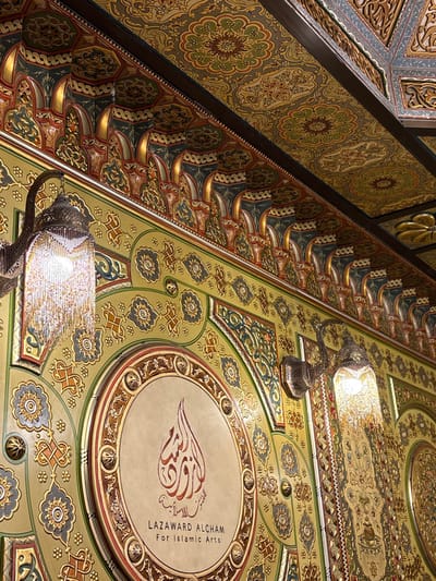 Ornate ceilings display intricate Islamic art patterns. Muted gold, green, and blue hues dominate the decorative motifs. Circular medallions feature Arabic calligraphy. Lamps with reflective surfaces hang, casting bright light on the artistry. The composition presents a rich visual tapestry, emphasizing the craftsmanship and design elements of traditional Islamic architecture. The words "LAZAWARD ALCHAM for Islamic Arts" are visible within one medallion.
