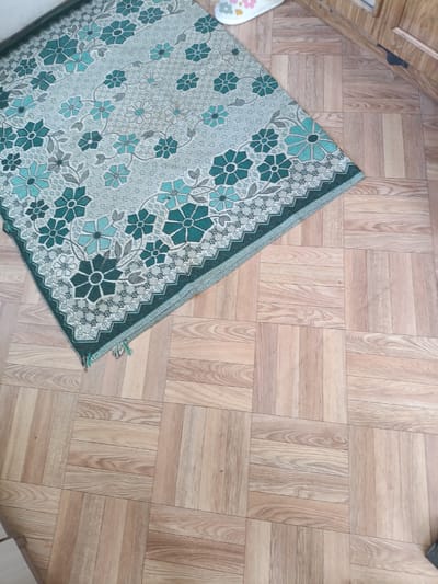 A rectangular, woven rug with a floral pattern is laid diagonally on a light brown, wood-patterned floor. The rug's pattern features light green and teal-colored flowers with dark green outlines and stems, set against a lighter, textured background. The rug has a dark green border with a zigzag pattern. A small white slipper with flower designs is visible at the top right corner of the rug. The floor is composed of square tiles arranged in a parquet pattern, with visible wood grain. To the right of the rug, a portion of a light brown wooden cabinet or furniture is visible. The scene appears to be indoors, likely in a residential setting, potentially in the village of Shipunovo, Russia, given the context. There are no people visible in the image.