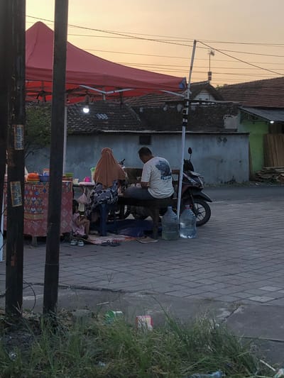 In Sentul, Indonesia, a street vendor stall is set up under a red canopy. The stall features a table covered with a patterned cloth, displaying various food items and containers. A person wearing an orange headscarf and a patterned outfit is seated at the stall, facing away from the camera. Next to them, another person in a light-colored t-shirt is also seated. A child in a pink dress is seated on the ground in front of the stall. In the background, houses with tiled roofs are visible, and a motorcycle is parked nearby. Two large water jugs are placed on the ground next to the seated individuals. The ground in front of the stall is paved with square tiles. The scene appears to be set during dusk, with a warm glow in the sky.