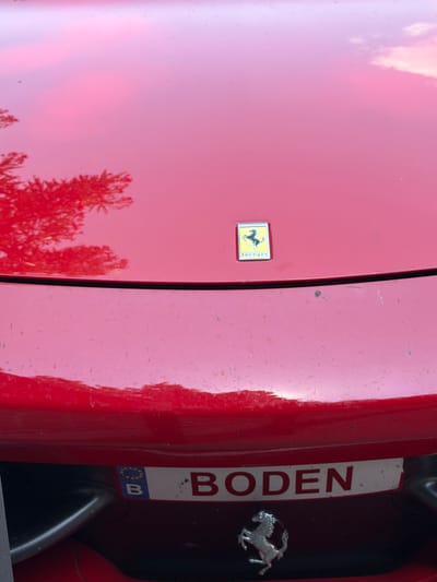This is a close-up, vertically oriented shot of a red Ferrari. The car's glossy paint reflects the sky and trees, suggesting it is outdoors during the daytime, possibly on a sunny day given the bright reflections. The image focuses on the front right side of the car, revealing a portion of the hood and a gap leading to the engine compartment.

On the left side of the image, the Ferrari's iconic prancing horse emblem is visible on a yellow and black background. To the right of the emblem, the word "ferrari" is etched vertically. Further to the right, a white plate with red lettering reads "BODEN". Below this plate, a smaller European Union license plate is partially visible, showing a blue background with stars and the letter "B". A silver prancing horse emblem, similar to the one on the hood, is also present on the right side of the frame. The overall condition of the car's paint appears to have some minor scuffs and dust, indicating it is a well-used vehicle rather than a pristine show car. The setting appears to be an outdoor location, with hints of a road or driveway implied by the reflections.
