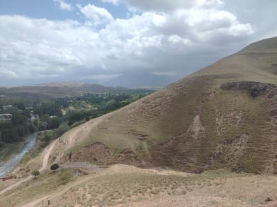 The image depicts a wide, panoramic view of a rural landscape in Tashbulak, Uzbekistan. In the foreground and midground, rolling hills are covered with dry, sparse vegetation and a winding dirt road. Erosion is evident on the slopes, with visible gullies and exposed soil. A river flows through the valley to the left, bordered by a dense line of trees and the edge of a village with buildings. In the background, more distant hills and mountains are visible under a partly cloudy sky. Two individuals, appearing small in scale due to the distance, are walking on the dirt road in the midground. The overall scene conveys a sense of arid, natural beauty with signs of human presence and habitation.