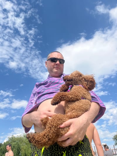 This media file shows a person holding a small, curly-haired brown dog. The person is wearing a purple and pink striped towel draped over their shoulders and black sunglasses. They are also wearing black swim trunks with a repeating yellow bunny logo and the word "PLAYBOY" in yellow letters. The dog appears to be wearing a colorful harness. The image is taken from a low angle, looking up at the sky, which is blue with scattered white clouds. The sun is bright, suggesting it is daytime. In the background, there are trees and other people, one of whom is looking down. The overall scene appears to be outdoors, possibly at a beach or park, given the swimwear and casual attire. The mood seems relaxed and summery. The media file appears to be a selfie.