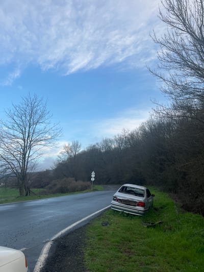 The image shows a silver car crashed off the side of a road in a rural area. The car is positioned on a grassy embankment, partially obscured by trees. The rear window of the car is shattered. The road is paved with a white line marking the edge. The sky is blue with scattered clouds. Trees line both sides of the road. A road sign is visible further down the road. The location appears to be Gabar, Bulgaria.