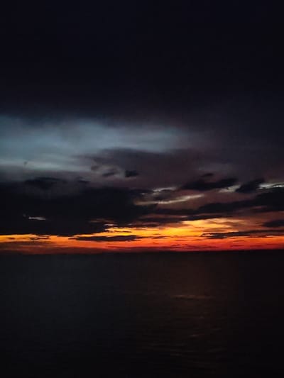 This is a vertically oriented, low-angle, wide shot of a sunset over the sea. The sky is a gradient of dark blue at the top, transitioning to grey and then to a vibrant band of orange and red near the horizon, where it meets the dark, calm sea. A few dark clouds are visible in the sky, interspersed with streaks of lighter color, suggesting the last rays of sunlight are breaking through. The sea is mostly dark and reflective, with a faint glimmer of light on the surface in the lower portion of the image. There are no people, animals, or man-made objects visible in the frame. The scene appears to be an undeveloped coastal area. The location is the city of Kvartsitnyi, Russia.