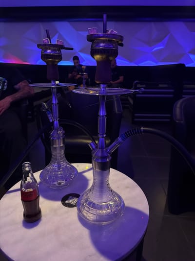 The image displays an indoor scene, likely a shisha lounge or bar in Gentbrugge, Belgium, characterized by dim, predominantly blue lighting. Two prominent hookahs are positioned on a white, circular, reflective table that occupies the right side of the frame.

Both hookahs feature clear, faceted glass bases, silver-toned metal stems, and dark hoses extending away from the table. Their top sections, made of a darker metal, hold bowls with glowing coals visible on top, indicating they are in use. Small, white cube-shaped coals can be seen on the top of the bowls.

On the table, near the lower hookah, rests a classic glass Coca-Cola bottle with its red label, partially filled with a dark liquid. A black, circular sticker with a QR code is also visible on the table, near the upper hookah. The table's glossy surface reflects the ambient blue light and elements of the hookahs.

In the blurred background, a wall with three-dimensional geometric patterns is illuminated by bright blue light, contributing to a modern, lounge-like atmosphere. Vague figures of people are discernible in the background, suggesting a social gathering. A person's arm and hand are visible in the lower-left foreground, near one of the hookahs.

The visible text includes "Coca-Cola" on the bottle's label. The overall mood is relaxed and social, indicative of an evening or nighttime event.