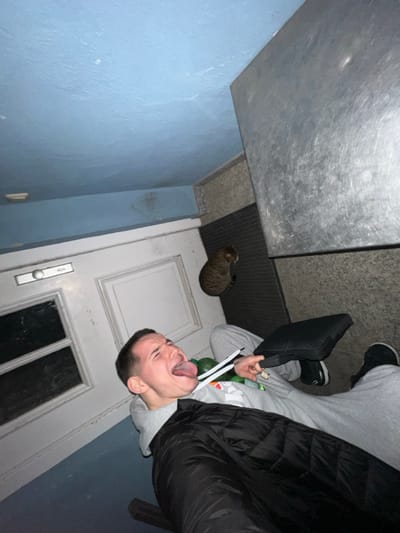 The image, captured in Benningen, Germany, displays a tilted interior scene featuring one male individual and one cat. A male with short, dark hair is depicted lying on his back on a light grey floor surface, positioned in the lower-left and center of the frame. He is wearing a black puffy jacket over a light grey hooded sweatshirt and grey sweatpants, with black sneakers on his feet. His mouth is wide open, with his tongue fully extended, and his eyes are squinted. His left arm extends towards the camera, suggesting he is holding the device. In his right hand, he holds a black rectangular object, possibly a small case. Near his upper torso, a green bottle and several white cylindrical items are present.

To the right of the man's head, a short-haired tabby cat with striped brown and grey fur is seated on a dark, ribbed doormat, facing to the right of the frame, away from the man. Behind the man, on the left side of the image, is a white door featuring two recessed rectangular panels and a horizontal silver handle assembly. The ceiling and an overhead structural beam are painted a light blue color, showing some discoloration and minor staining. A large, dull, metallic surface, appearing scuffed and scratched, occupies the upper-right quadrant of the image, extending downward towards the floor level. The environment is lit dimly and evenly.