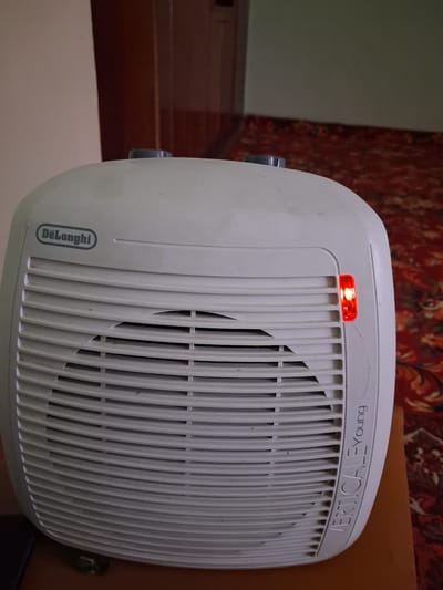 A white DeLonghi brand fan heater, with a red indicator light illuminated on its right side, is positioned on a wooden surface in what appears to be a residential setting. The fan grille is visible, with some minor dust accumulation and a small imperfection in the plastic. The background consists of a patterned carpet in shades of red and orange, and a wall that is partially visible. The location is identified as Kahramon, Uzbekistan.