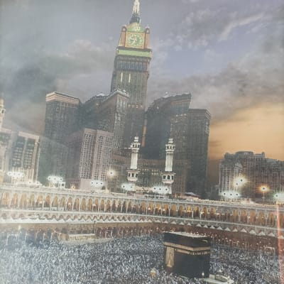 The image depicts an aerial or elevated view of the Masjid al-Haram complex in Mecca, Saudi Arabia. In the foreground, a massive congregation of individuals, appearing as pilgrims, are densely arranged while performing circumambulation (Tawaf) around the Kaaba, a prominent cuboid structure draped in black cloth with gold detailing, centrally positioned in an open courtyard. Surrounding the courtyard are the multi-tiered structures of the Grand Mosque, characterized by numerous arched walkways and supporting columns. Two tall, white minarets with multiple balconies are visible in the midground, rising above the mosque's lower levels. Dominating the background is the Abraj Al-Bait Clock Tower, a towering skyscraper with a large, illuminated clock face and a spire topped with a crescent. Numerous other tall, dark-hued skyscrapers surround the complex. The sky displays a transition from a lighter, possibly overcast grey on the left to an orange-yellow hue on the right, suggesting either dusk or dawn, with visible cloud formations. Artificial lights are discernible within the mosque structure and on the surrounding buildings. No specific individual interactions are clearly visible due to the vast scale and crowd density, but the collective action of circumambulation is evident.