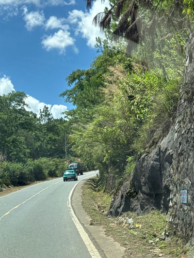 This is a photograph taken from a moving vehicle on a paved road that curves to the left, bordered by a steep, rocky incline on the right and lush vegetation on the left. The road has yellow and white dividing lines. In the foreground, a teal-colored car is driving away from the viewer, followed by a dark-colored truck. The environment is characterized by dense green foliage and trees that line the roadside, extending up the slopes. The sky above is bright blue with scattered white clouds. There is some trash visible at the base of the rocky incline on the right side of the road. On the rocky face, there is a small, light blue sign with some text on it. The scene appears to be a scenic mountain road in a rural or natural area. This image depicts a road in Thôn 6, Vietnam.