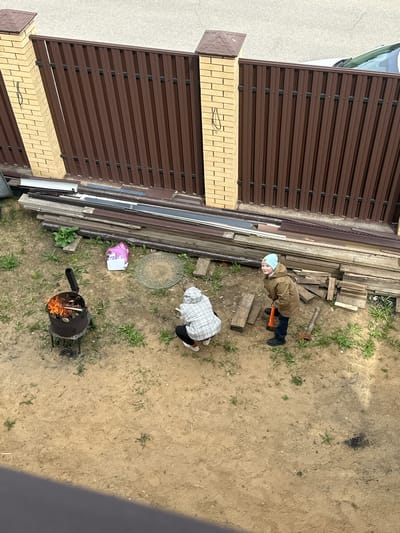 The image shows an overhead view of a backyard area, presumably in слобода Старая, Russia. A section of brown metal fence with a brick base runs along the left edge of the frame. Adjacent to the fence are stacks of lumber and various building materials. Two people are visible: one wearing a brown jacket and a light blue head covering is standing and holding an orange-handled tool. The second person, wearing a white and gray patterned head covering, is bending over. A black metal grill or incinerator containing burning wood is positioned in the lower part of the frame. The ground is mostly dirt with patches of grass. A plastic bag and a round manhole cover are near the lumber pile.