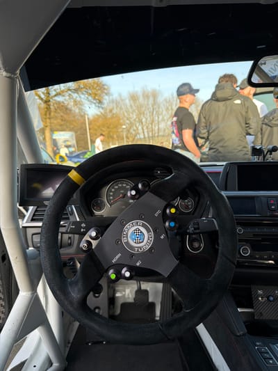 The image provides an interior view from a customized vehicle, looking out through the windshield onto an outdoor gathering in Meuspath, Germany. In the foreground, a black suede-wrapped racing steering wheel with a flat bottom is centered. Its hub features a black metallic plate with "BIMMER TUNING" and a circular logo displaying a blue and white checkered pattern. Multiple function buttons are affixed to the steering wheel, including a yellow button labeled "DTC," a blue button labeled "M1," a green button labeled "HORN," and a white button labeled "M2."
Behind the steering wheel, a dashboard is visible, housing an analog instrument cluster displaying a speedometer and tachometer. A small black digital display unit is mounted to the left of the steering wheel. A white tubular roll cage structure is prominent on the vehicle's left side. The vehicle's interior appears modified for performance, with dark trim and visible carbon fiber accents on the center console.
Beyond the windshield, the outdoor scene is illuminated by natural light under a clear blue sky. Several individuals are present on a paved surface. In the mid-ground, one person wears a black baseball cap and a dark t-shirt with a colorful graphic on the back. Another individual stands with their back to the camera, wearing a dark jacket bearing "The North Face" branding. Other people are visible in the background. Deciduous trees with bare branches are present, along with a light pole and indications of other vehicles and signage in the distance. The setting suggests an automotive event or gathering.