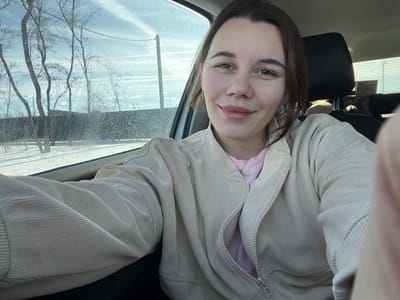 The image depicts a female subject seated inside a vehicle, located in the city of Shchepkin, Russia. The subject has brown hair and a light complexion, looking directly at the camera with a slight smile. She is wearing a light beige, corduroy-textured jacket, partially unzipped to reveal a pink garment underneath, and a thin silver-colored necklace. The subject's right fingers are visible on the far right edge of the frame, indicating a self-portrait. Through the vehicle's driver's side window, which appears dirty or streaked, a winter landscape is visible. This exterior scene includes snow-covered ground, several bare deciduous trees, a dark-colored horizontal fence in the middle ground, and a tall utility pole on the right. The sky is light blue with multiple white contrails.