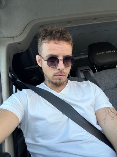 The image shows a young man sitting in the driver's seat of a vehicle, with a seatbelt fastened across his chest. He is wearing a white t-shirt and sunglasses. The interior of the vehicle is visible, including the headrest of the seat behind him, which has the word "DUCATO" embossed on it. The man has short, spiky brown hair and appears to have tattoos on his neck and arm. The lighting suggests it is daytime, and the overall scene is consistent with being inside a car. There is no information in the image that can identify the location as Mikhmoret, Israel.