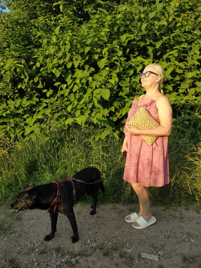 A woman and a black dog are walking on a gravel path. The woman is wearing a pink patterned dress, white slides, and glasses. She is holding a woven clutch. The dog is wearing a red harness. Behind them is a dense green bush and tall grass. The sun is shining, casting shadows, suggesting it is either morning or late afternoon. The setting appears to be a natural outdoor area, possibly a park or recreational spot. The overall mood is relaxed and peaceful. No visible text is present in the image.