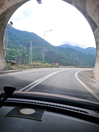The image shows a view from inside a vehicle exiting a tunnel and approaching a curved road with mountains in the background. The road has double white lines. The tunnel's concrete arch is visible on the left and right sides of the frame. Outside the tunnel, there are green, tree-covered mountains under a cloudy sky. A power line tower and street lights are visible along the roadside. The dashboard of the vehicle, including a suction cup mount and a decorative mat with an image of sunglasses, is in the foreground. The location is implied to be a road near Artvin, Turkey, given the mountainous terrain and road infrastructure.