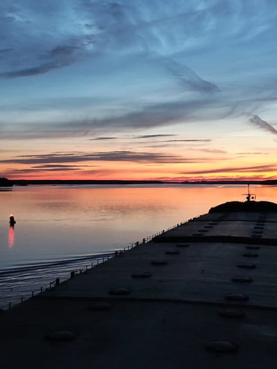 This image depicts a sunset over a body of water, likely a bay or sea, with the foreground dominated by the deck of a large vessel. The sky is a gradient of blues, grays, and oranges, with streaks of clouds indicating movement and depth. The sun, low on the horizon, casts a warm, orange glow across the water, reflecting the vibrant colors of the sky. A navigational buoy with a red light is visible in the mid-ground, marking a channel or hazard. The deck of the vessel in the foreground is weathered and utilitarian, with visible hatches or access points. The overall impression is one of tranquility and the vastness of nature. The location is stated to be Береговой, Russia.
