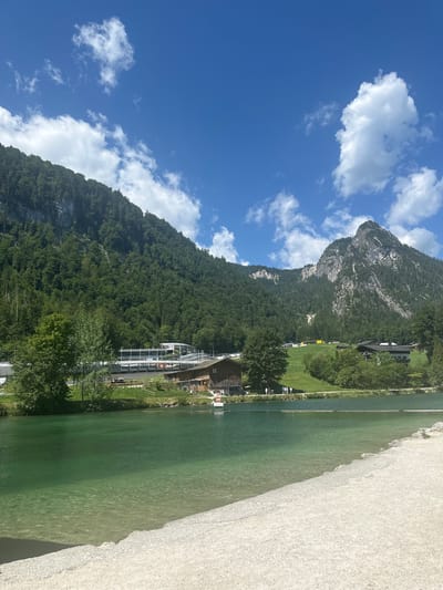 This is a vertical, eye-level, outdoor shot of a scenic lake and mountain landscape. The foreground is dominated by a sandy embankment that slopes down to the clear, turquoise water of the Königssee lake. The water is shallow near the shore, revealing the pebbled lakebed. The midground features the lake extending into the distance, with a small white boat visible in the water.
On the far bank of the lake, there is a green grassy area with scattered trees. Beyond the trees, several buildings are visible, including a large, modern-looking complex with white and gray structures, possibly a visitor center or recreational facility, and traditional wooden houses with dark roofs. Further up the slope, more buildings are nestled among the trees.
The background is dominated by a large, verdant mountain, densely covered with evergreen trees. The mountain's rocky peak is visible against a bright blue sky filled with scattered white, fluffy clouds. The overall impression is one of natural beauty and a recreational area. The location is Schönau am Königssee, Germany, given the iconic landscape. There are several people visible in small groups along the shore of the lake and near the buildings.