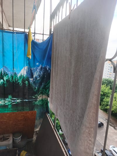 The image depicts a cluttered balcony in what appears to be a residential building in Sof'ino, Russia. The scene is characterized by laundry drying on a clothesline. A large, light-colored towel is prominently displayed, held by clothespins to a thin, reddish-orange line. To the left, a blue tarp with a scenic mountain and lake landscape is partially visible, along with a small, yellow garment also hung on the line.
The balcony itself is framed by what looks like metal bars, suggesting it's an outdoor space. Below the clothesline and tarp, there are various objects, including a piece of plywood, several buckets or containers, and what seems to be some construction debris.
In the background, through the gaps in the balcony structure, a residential building with multiple floors and windows is visible. Lush green trees surround the area, indicating a setting with some vegetation. The sky appears overcast or hazy, suggesting a cloudy day. The overall impression is one of a functional, somewhat untidy living space, where everyday activities like drying clothes are taking place. No people are visible in the frame. The weather appears to be temperate, suitable for drying clothes outdoors.