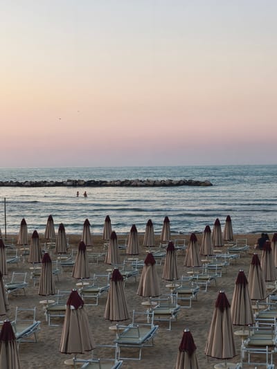 The image depicts a beach scene at dawn or dusk, with the sky exhibiting a soft gradient of pastel colors from light blue at the top to pink and lavender near the horizon. In the foreground, numerous closed beach umbrellas and empty lounge chairs are arranged in neat rows on the sand. The umbrellas are a neutral beige color with dark red accents. The sea is calm with gentle waves, and a breakwater made of large rocks is visible in the middle ground. Two people are standing in the shallow water near the breakwater. In the far distance, a few birds are visible in the sky. The setting is characteristic of the San Salvo Marina beach, a popular tourist destination in San Salvo, Italy.