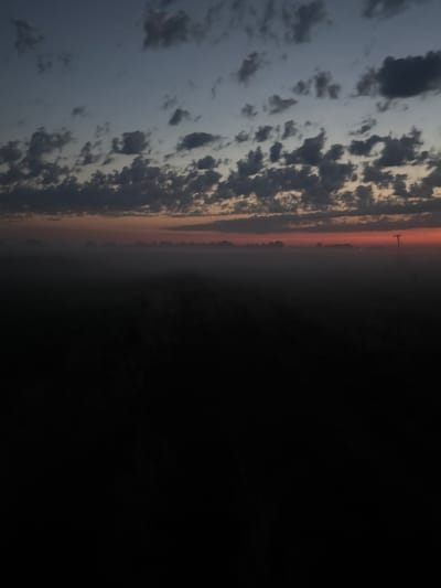 The image captures a foggy landscape at dawn or dusk, likely in the city of Ворошиловка, Kazakhstan. The sky displays a gradient of colors, transitioning from a deep blue at the top to shades of gray and orange near the horizon. Scattered clouds are visible in the upper portion of the sky. The middle ground is obscured by a thick layer of fog, which obscures any details of the terrain or any structures that might be present. A solitary pole, possibly a utility pole or a lamppost, stands out against the hazy horizon on the right side of the image. The foreground is extremely dark, rendering it difficult to discern any features. The overall impression is one of a quiet, atmospheric, and somewhat desolate scene.