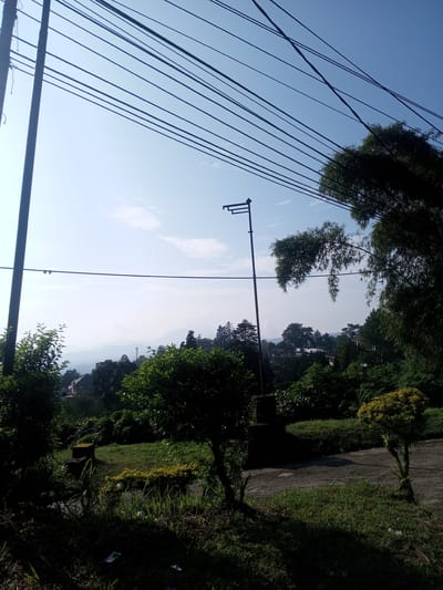 The image depicts a daytime scene in Phouviang-Gnai, Laos. Overhead power lines dominate the top of the frame against a pale blue sky with a few scattered clouds. A tall, dark metal post with an apparatus at the top stands prominently. The midground features lush green vegetation, including various trees and bushes. In the background, there is a hilly landscape with buildings and possibly a cell tower visible amidst the trees. A paved path curves through the foreground with grass and small plants growing alongside it.