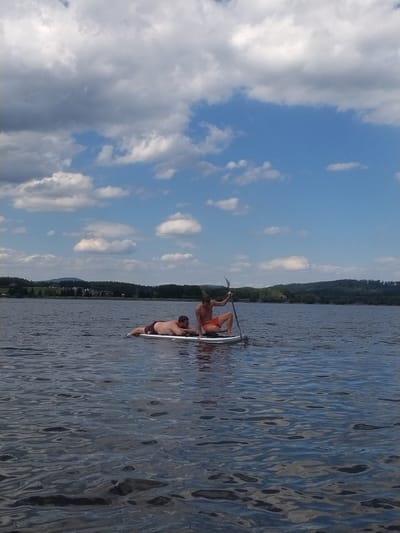 Two adult males are on a paddleboard in a body of water, likely a lake, under a cloudy blue sky. One male is lying on his stomach on the front of the paddleboard, wearing red swim trunks. The second male is kneeling on the back of the paddleboard, holding a paddle and wearing orange swim trunks. The background shows a shoreline with trees and distant hills. The water is rippled. The scene appears to be outdoors in a natural setting. The location is identified as Radslav, Czechia.