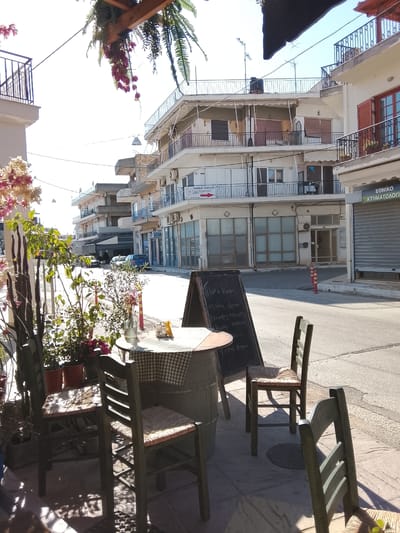 This image, captured in Λεχαινά, Greece, depicts a quiet street scene featuring a small outdoor café setup. The focal point is a round table resting on a barrel, covered with a checkered cloth. The table is set with a glass bottle, possibly holding water, a container with utensils, and a small yellow item. Three green wooden chairs with woven seats surround the table. Next to the table stands a chalkboard menu displaying items in Greek script.

The street is largely deserted, with white buildings on either side, some featuring balconies. The architectural style suggests a Mediterranean setting. The building on the right has a closed metal shutter with the word "ΕΘΝΙΚΟ ΚΤΗΜΑΤΟΛΟΓΙΟ" on it. Further down the street, a couple of parked cars are visible. Overhead, wires stretch across the street, connecting the buildings. The lighting suggests it is a sunny day. The presence of potted plants and flowers around the café seating adds a touch of nature to the scene.