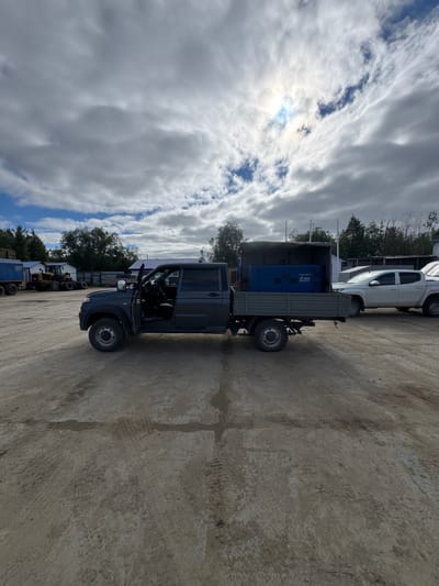 The image shows a dark grey SUV parked on a concrete lot with a slight upward slope. The SUV has its driver's side door open. Behind the SUV, a flatbed truck is parked, loaded with a large blue generator. A white pickup truck is parked in front of the flatbed truck. The scene is outdoors, with trees and a building visible in the background. The sky is cloudy, with patches of blue sky and sunlight breaking through. The time of day appears to be daytime. There is visible text on the blue generator that reads "GENTEC" and "WWW.GENTEC.RU". The setting appears to be an industrial or storage area. The image is taken in Parma, Russia.