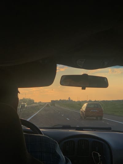 The image captures a view from inside a vehicle traveling on a highway during sunset. The perspective is from the driver's seat, with a portion of the steering wheel and dashboard visible in the foreground. A person's shoulder and head are partially visible on the left side, suggesting the driver or a passenger is in the frame. In the middle ground, other vehicles are seen on the road, including a reddish-orange car in the right lane. The sky is illuminated by the setting sun, casting a warm, orange glow across the scene and creating a hazy atmosphere. The landscape consists of flat terrain with some vegetation and structures, including what appears to be a small building or tower in the distance on the left. The overall impression is of a journey on a rural highway in Belarus at dusk.