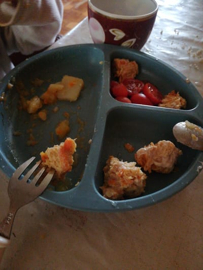 The image shows a divided blue plate with food remnants, a fork, and a spoon on a table covered with a patterned tablecloth. The plate contains pieces of what appears to be a casserole or ground meat dish with visible shredded carrots, as well as some sliced tomatoes. A fork is positioned on the left side of the plate, with a piece of food impaled on it. A spoon with a dollop of food is visible in the upper right section of the plate. In the background, a portion of a person's torso wearing a white garment is visible, and a red and white patterned bowl is also present. The lighting suggests it is daytime. The food appears to be partially eaten, indicating a meal in progress. The overall scene is casual and domestic. No legible text is visible in the image. The location context is Dubrovka, Russia.