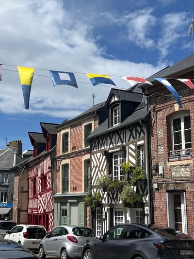The image depicts a street scene in Honfleur, France, with a row of traditional half-timbered buildings lining the right side. The architecture features a mix of exposed wooden beams against white plaster and red brick facades, with steeply pitched dark roofs. Several windows are visible, some with black ironwork. A string of nautical signal flags, including yellow, blue, and red patterns, stretches across the foreground against a bright blue sky with scattered white clouds. In front of the buildings, several cars are parked or in motion. To the left, a white car and a silver hatchback are prominent, with a dark-colored Tesla visible in the immediate foreground on the right. The street appears to be paved. The overall impression is one of a charming, historic European town on a clear day.